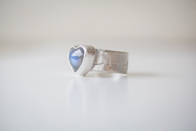 A sterling silver ring featuring a heart shaped labradorite gemstone with a bright blue flash. The band is hand sawed with an EKG heart rhythm. 
