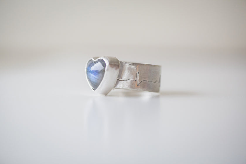 A sterling silver ring featuring a heart shaped labradorite gemstone with a bright blue flash. The band is hand sawed with an EKG heart rhythm. 