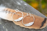 Silver feather-shaped earrings on a wooden surface with a blurred natural background