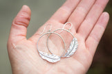 Silver hoop earrings with feather designs held in a hand against a blurred background