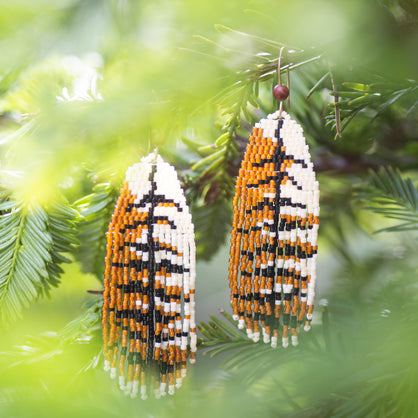 Beaded earrings that look like Red Tailed Hawk feathers hanging on a branch with green leaves in the background
