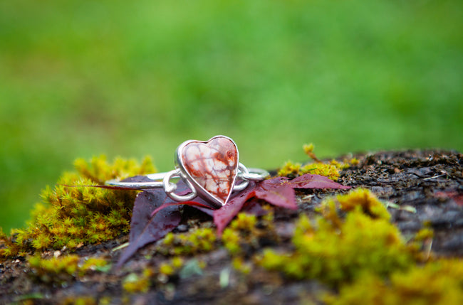 A sterling silver bracelet with a large heart shaped rhyolite gemstone. Set against a green natural background. 