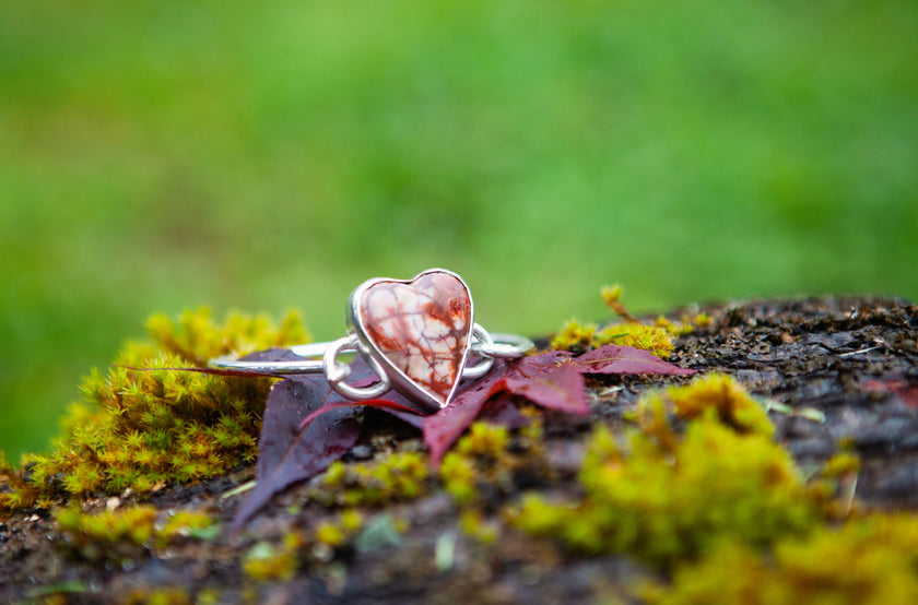 A sterling silver bracelet with a large heart shaped rhyolite gemstone. Set against a green natural background. 