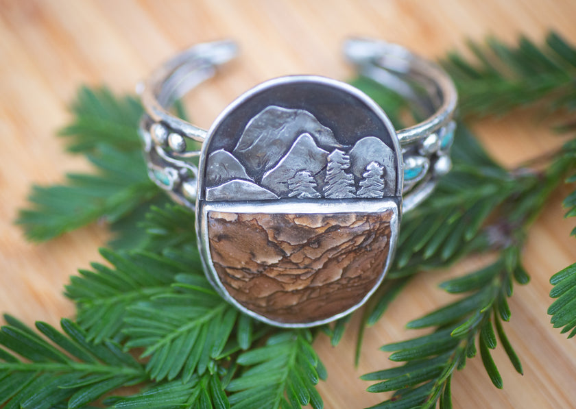 Handmade sterling silver cuff featuring a large mountain scene with pine trees and a scenic Deschutes jasper as well as small turquoise stones. 