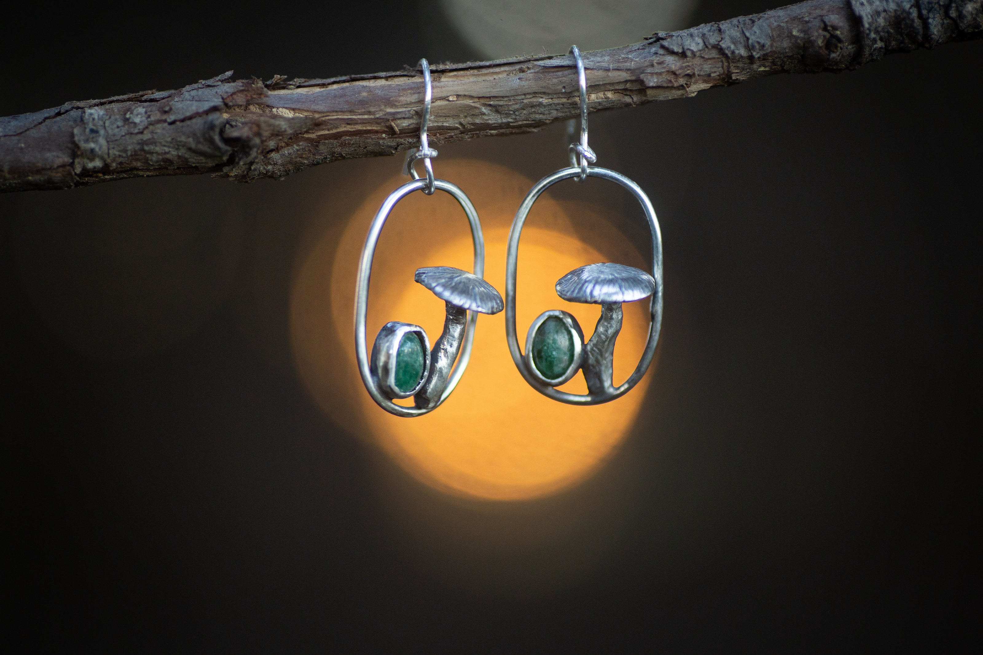 These detailed handmade earrings feature whimsical mushrooms and tiny oval aventurine gemstones. They are pictured against a lit forest background. 