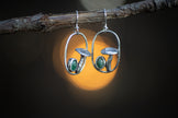 These detailed handmade earrings feature whimsical mushrooms and tiny oval aventurine gemstones. They are pictured against a lit forest background. 